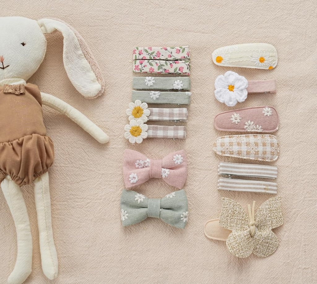 A collection of hair clips arranged neatly, featuring floral designs and bows, next to a soft toy rabbit on a textured light background.