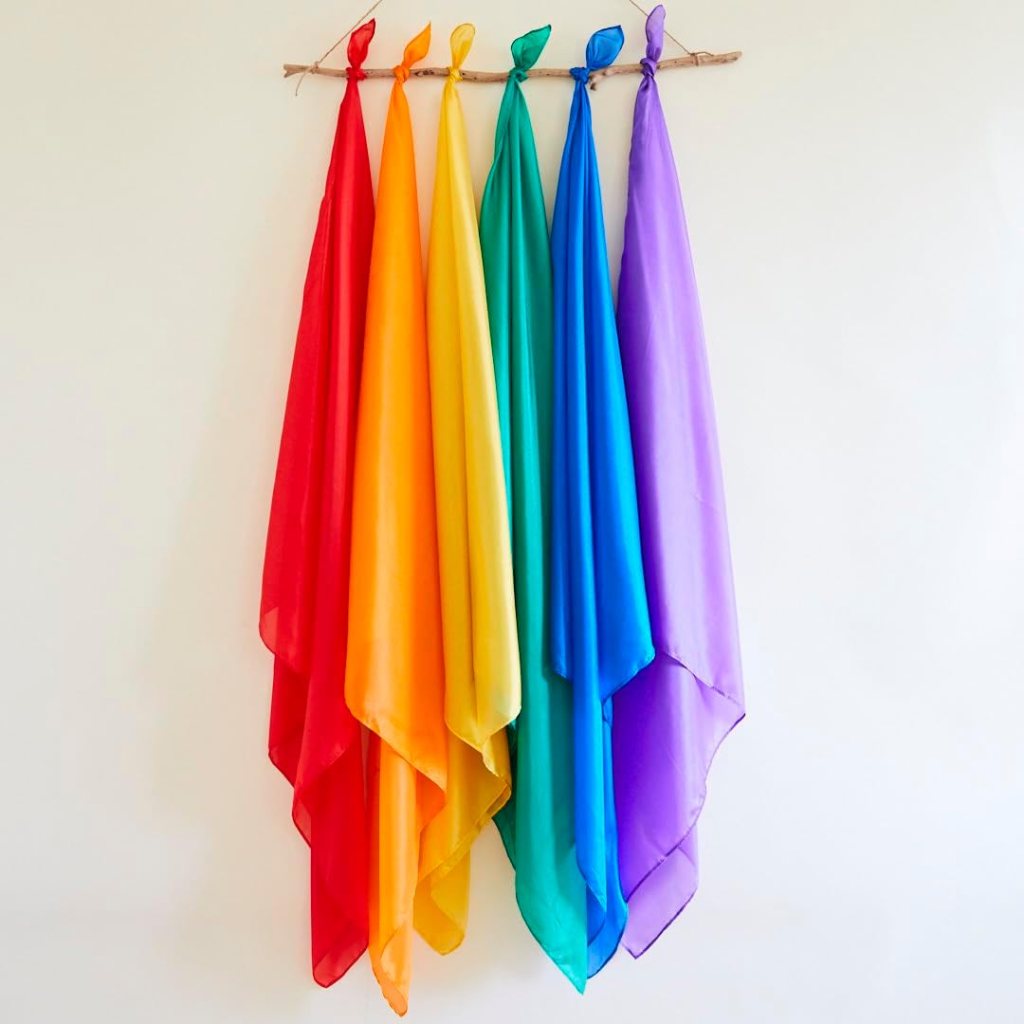 A display of six colorful scarves hanging from a wooden rod against a neutral wall, with colors ranging from red to purple.